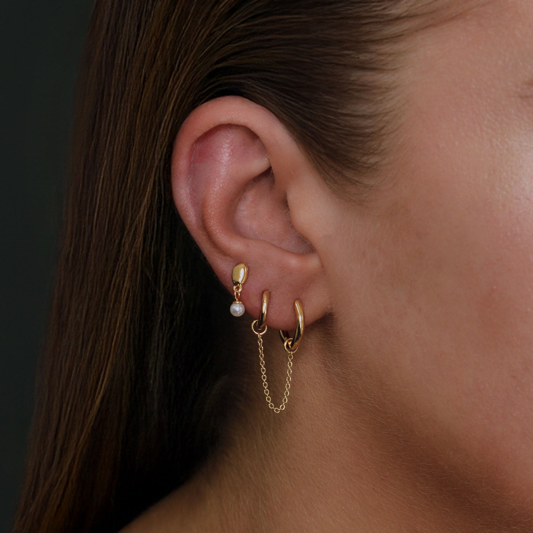 A close-up of a person's ear wearing multiple gold earrings, including hoop earrings with a dangling chain, a small pearl stud, and a gold dome earring, against a dark background.
