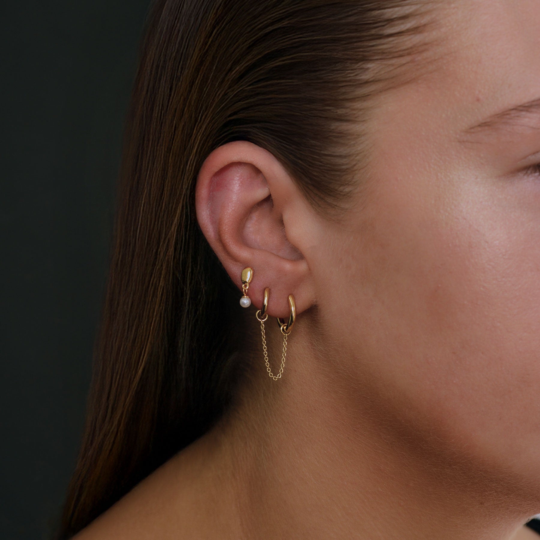 A close-up of a person's ear wearing a stack of gold earrings, including a small pearl stud, a hoop with a dangling chain, and a gold huggie, against a dark background.