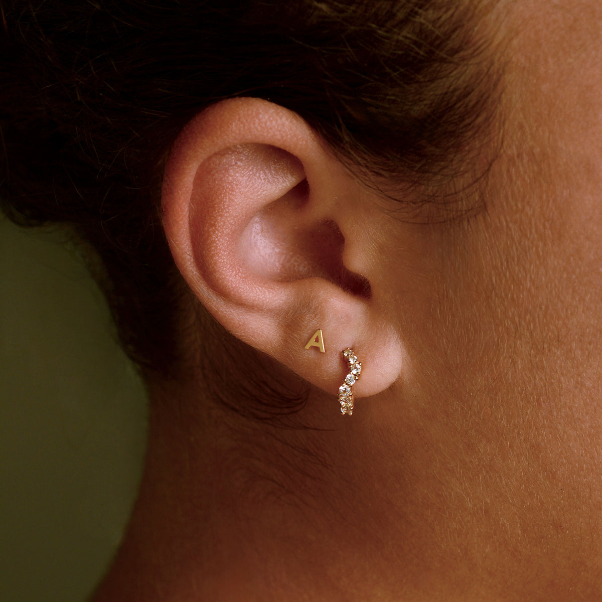 Gold A letter stud earring with a curved white gem set hoop earring on a models ear.