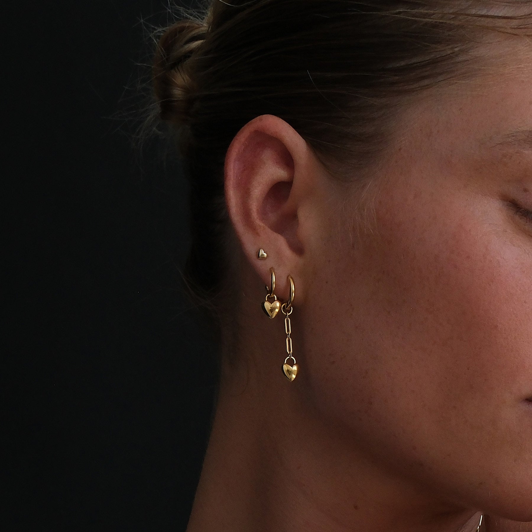 Model wearing a gold plated earring stack with a heart stud, heart hoop and a heart chain drop huggie earring.