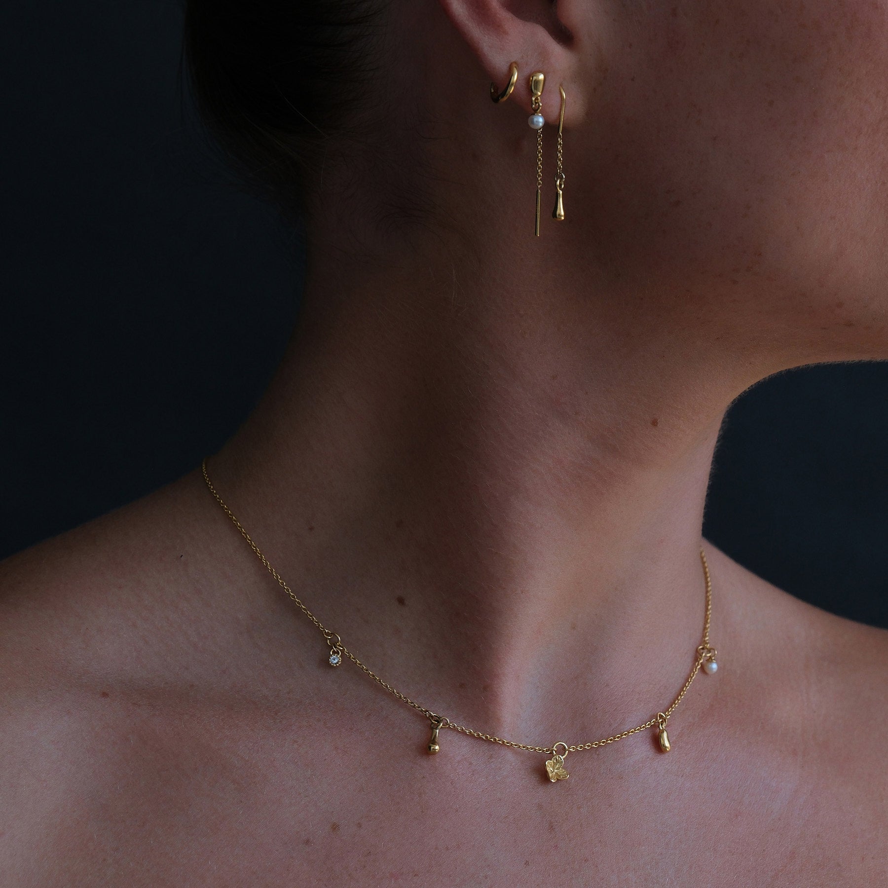 Close up of model wearing an 18k gold vermeil charm necklace featuring a flower, pearl and organic-shaped charms. Model is wearing gold plated pearl drop earrings with a mini huggie. 