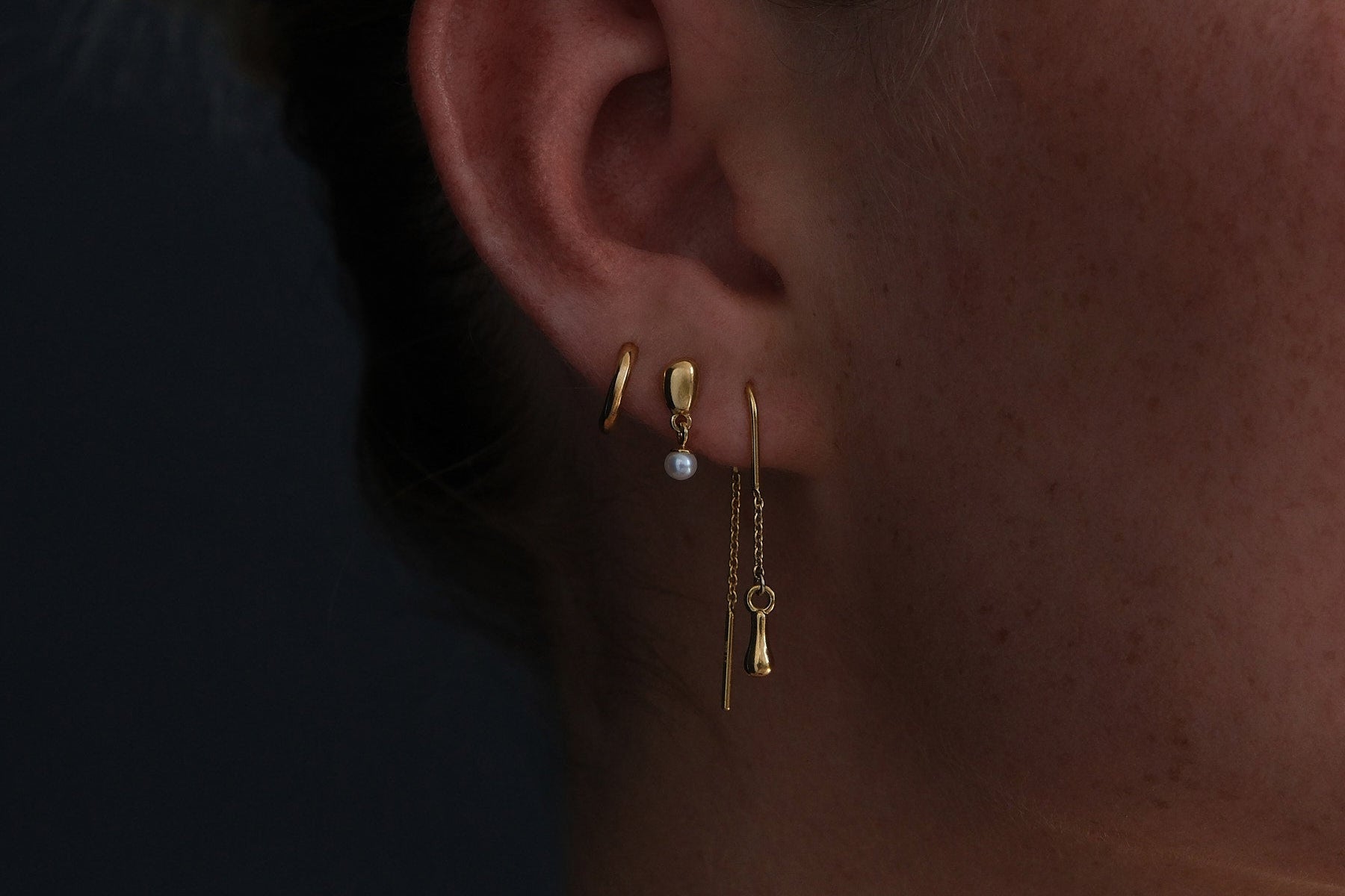 Close up of gold plated earring stack featuring mini huggie, pearl drop stud and a drop thread earring.