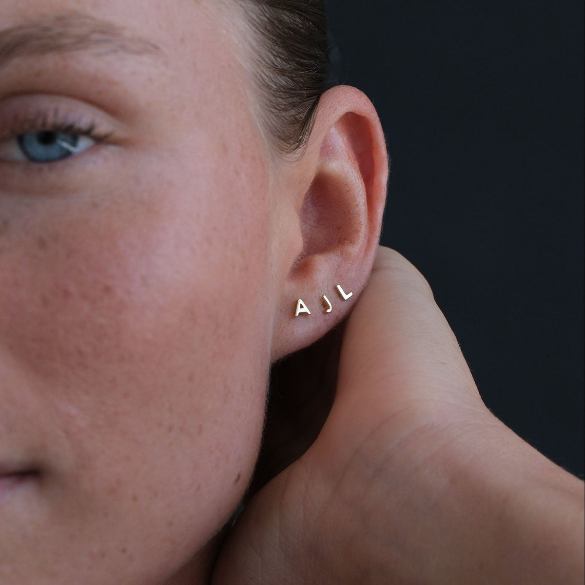 Close-up of a person wearing a gold earring with 'A.J.' on a dark background